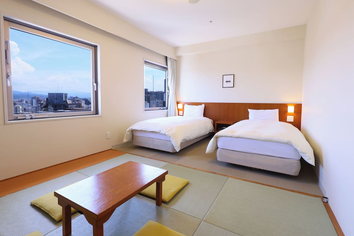 A hotel room with two beds, a wooden table, tatami mats, and a city view.