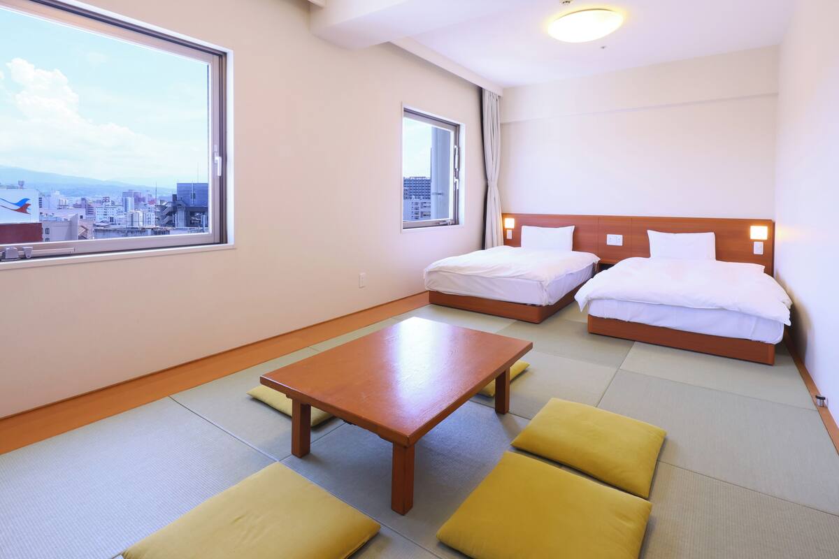A hotel room with tatami flooring, a wooden table, two beds with white bedding, and a city view through large windows.