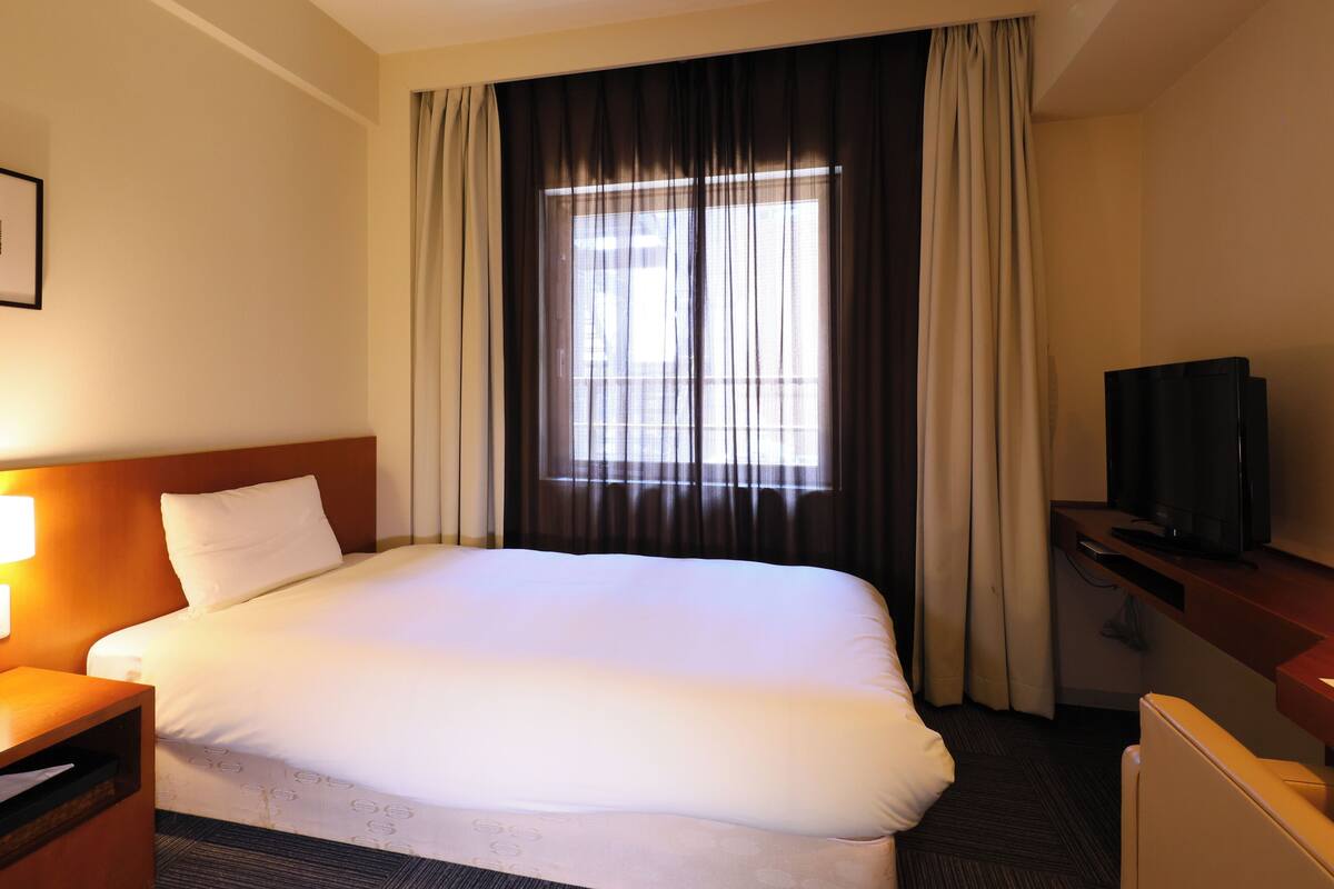 A hotel room with a large bed, a desk with a lamp, a television, and a window with curtains.