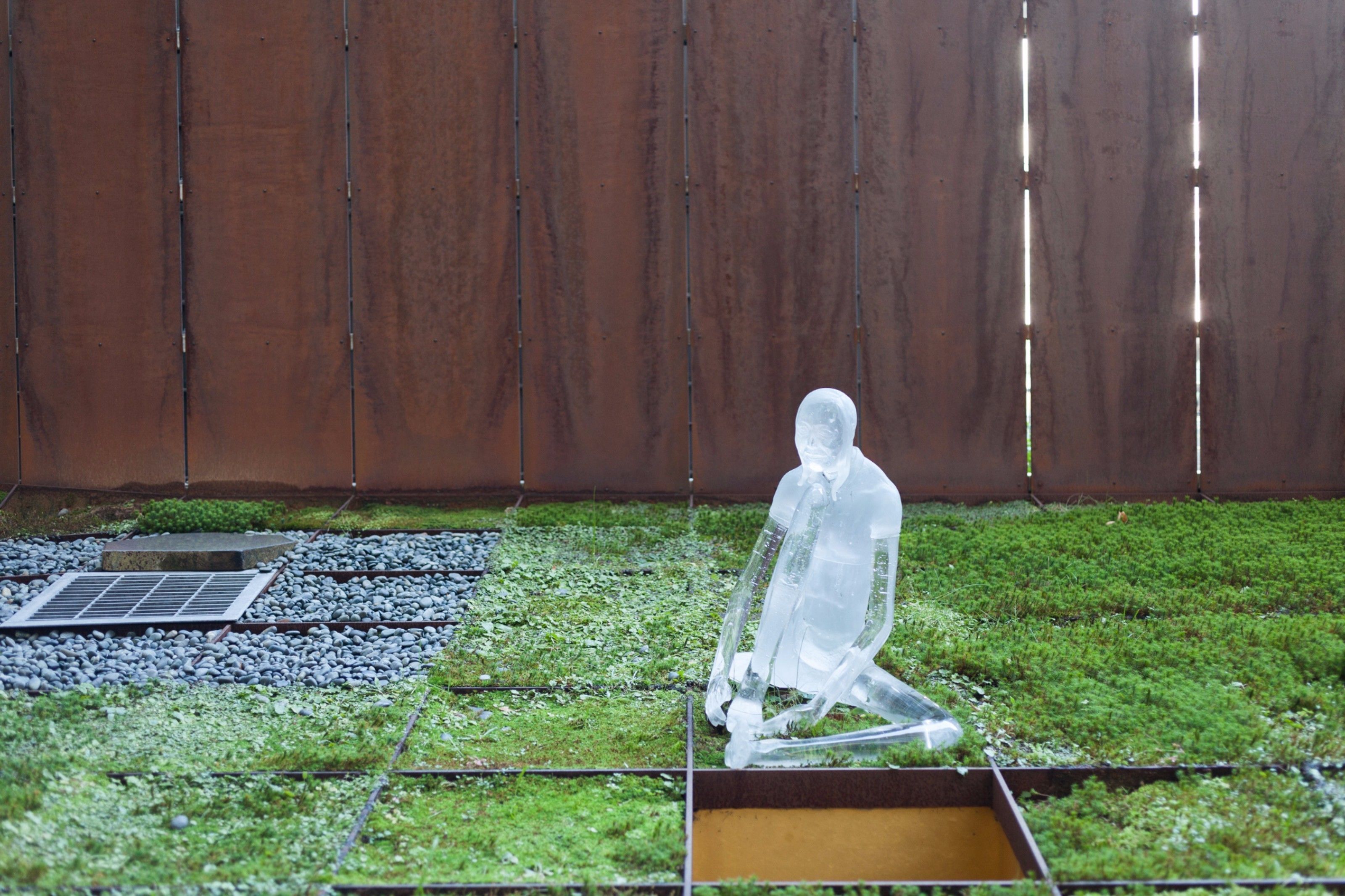 A transparent sculpture on a grassy area with a metal grate and a wooden fence in the background.