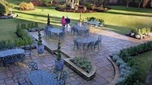 A patio area with stone flooring, metal tables, and chairs, surrounded by landscaped gardens and a lawn. - The Collingwood Arms Hotel (Cornhill on Tweed)