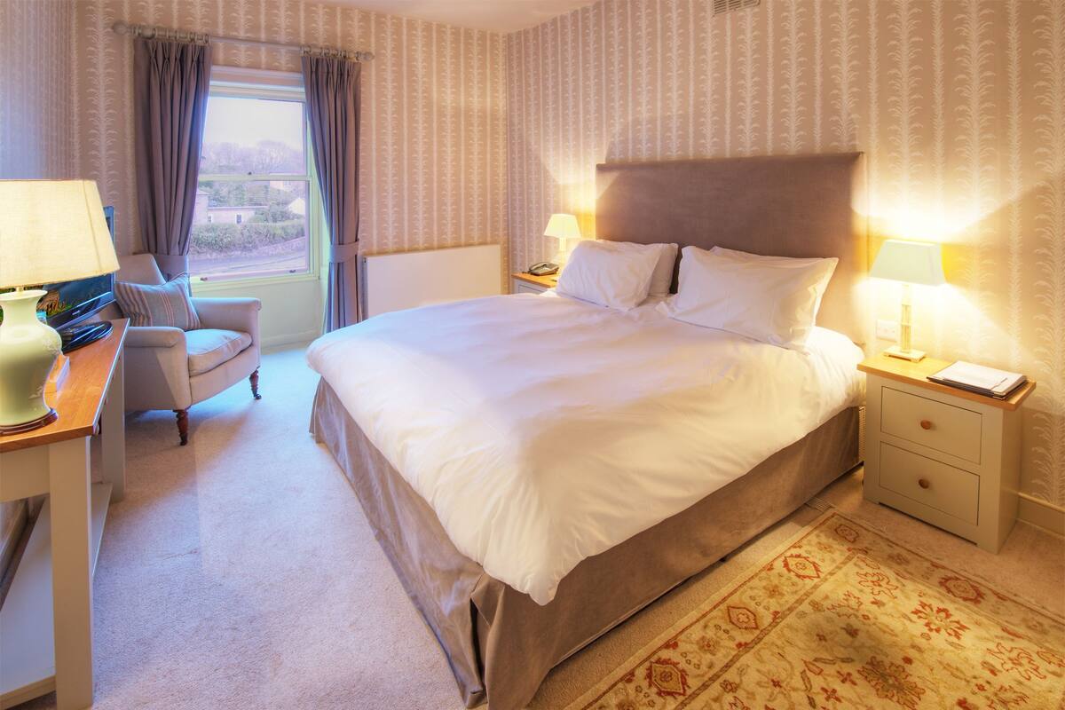 A hotel room with a large bed, a desk with a lamp, a chair, and a view of the outdoors through a window.
