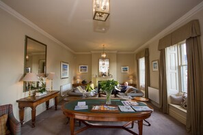 A spacious living room with a round wooden table, comfortable seating, a fireplace, and large windows with curtains.
