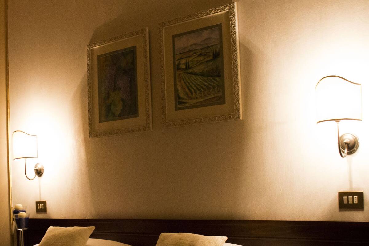 Hotellihuone, jossa on sänky, kaksi seinään kiinnitettyä lamppua ja kaksi kehystettyä kuvaa seinällä.