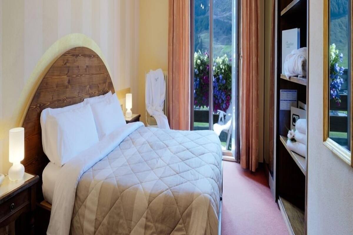 A hotel room with a large bed, a wooden headboard, a nightstand with a lamp, a robe, a wardrobe, and a view of a garden through a sliding door.