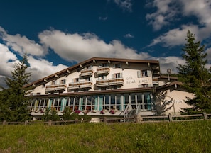 A hotel with multiple balconies, a ski slope in the background, and a clear sky.