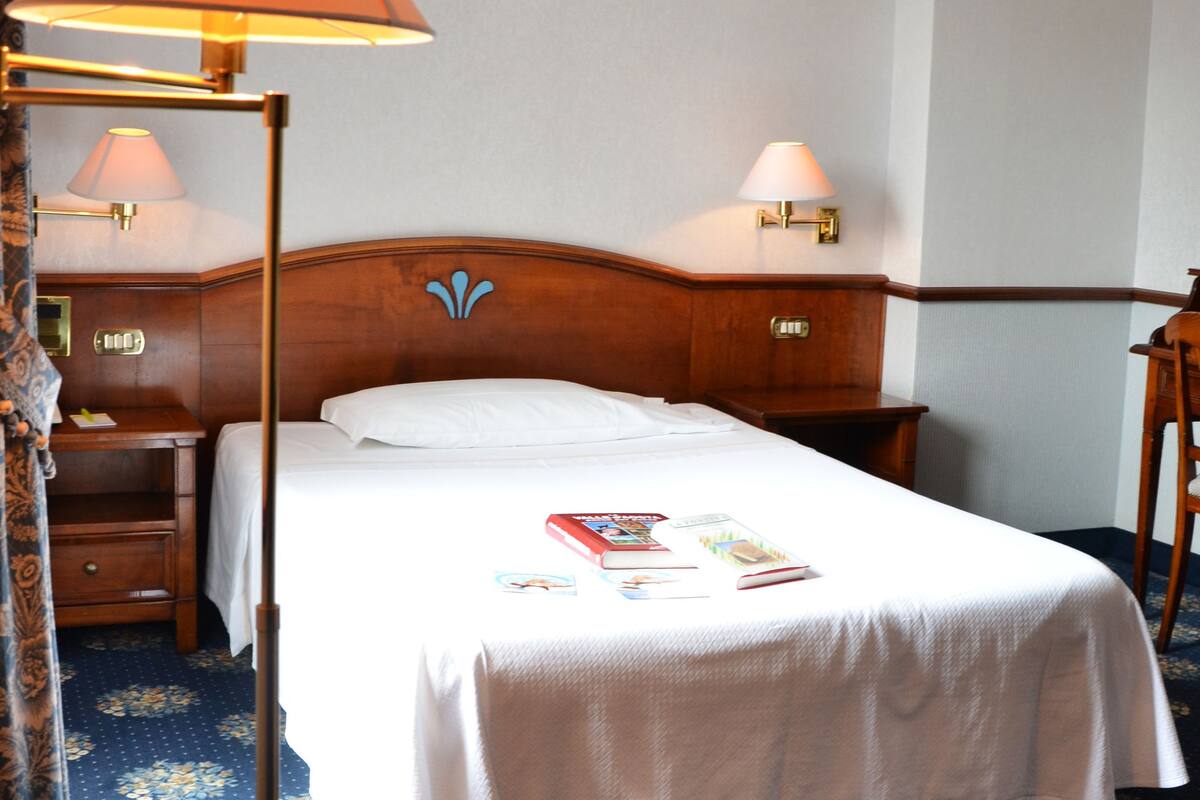 Una habitación de hotel con una cama bien hecha, una mesita de noche con una lámpara, un cabecero de madera y una alfombra estampada con motivos florales.
