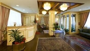 A spacious living area with a large rug, a sofa, and a coffee table. There is a plant in a gold pot and a chandelier hanging from the ceiling. - Hotel De la Ville (Saint-Vincent)