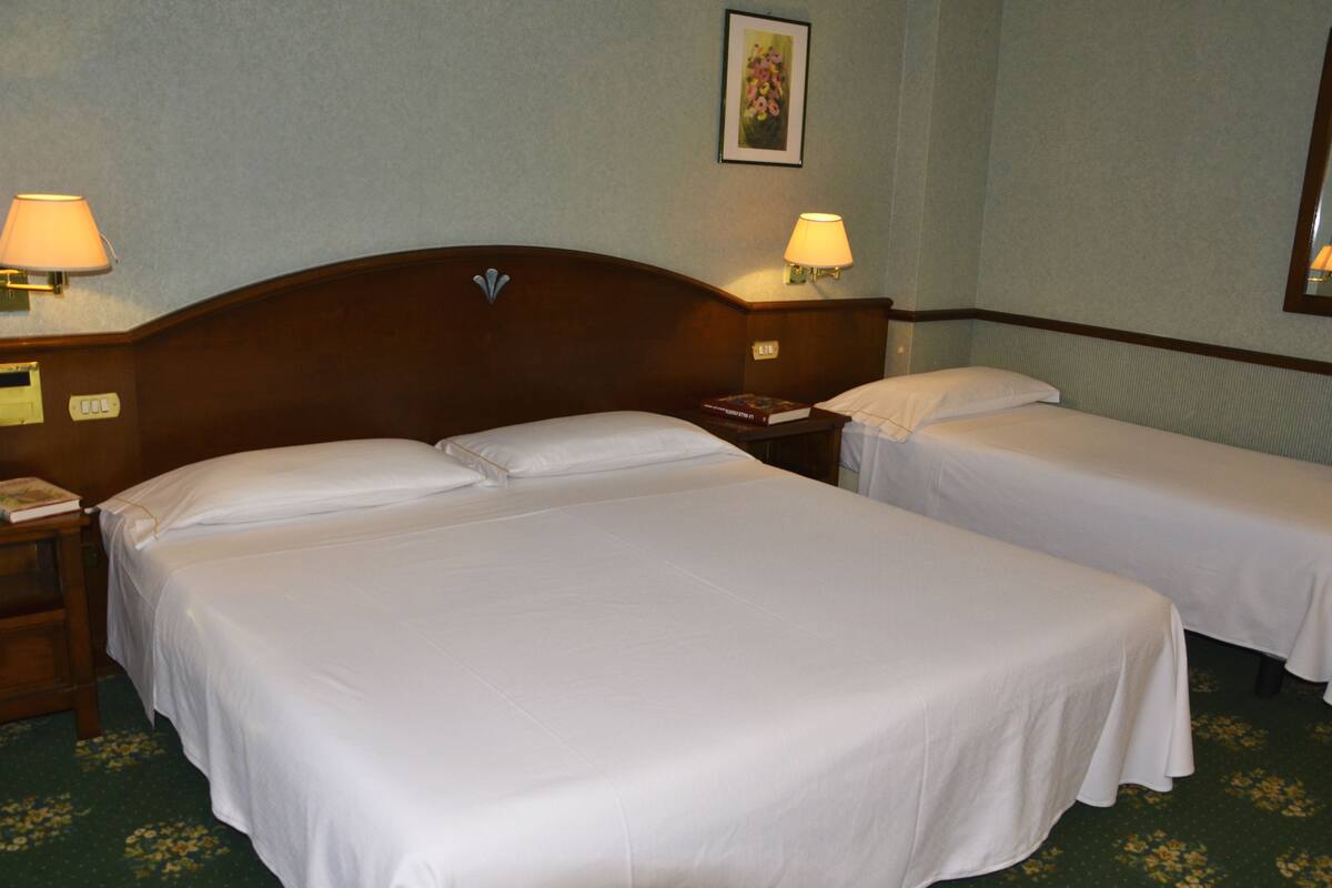 Habitación de hotel con dos camas individuales, una mesita de noche con lámpara, una mesita con un libro y un cuadro en la pared.