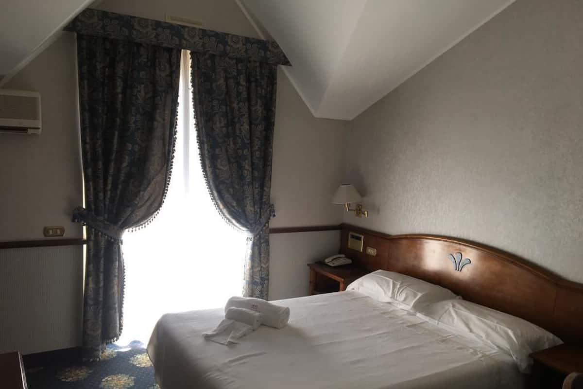 Una habitación de hotel con una cama bien hecha, una mesita de noche con lámpara y una ventana con cortinas.