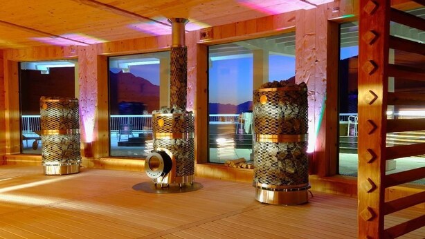 A room with wooden walls and large windows, featuring three metallic stoves with a textured design. - Hotel De la Ville (Saint-Vincent)