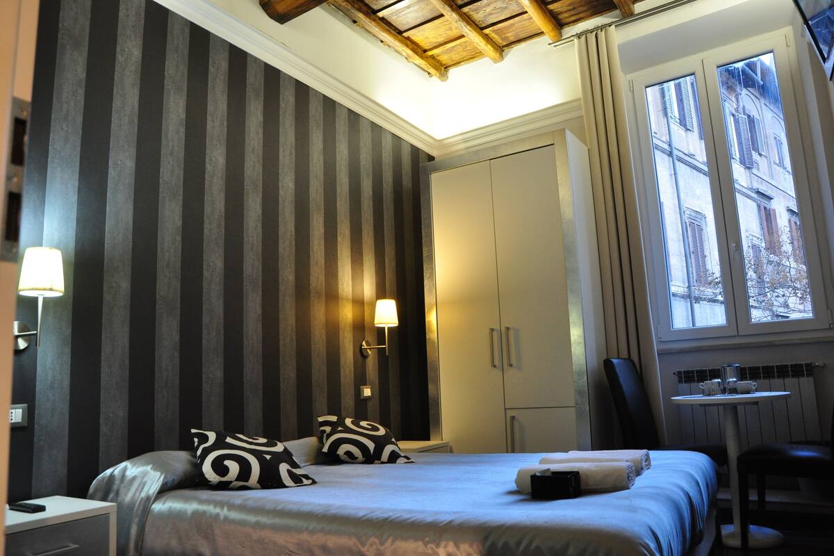 A hotel room with a bed, bedside lamps, a desk, and a window with a view of buildings.