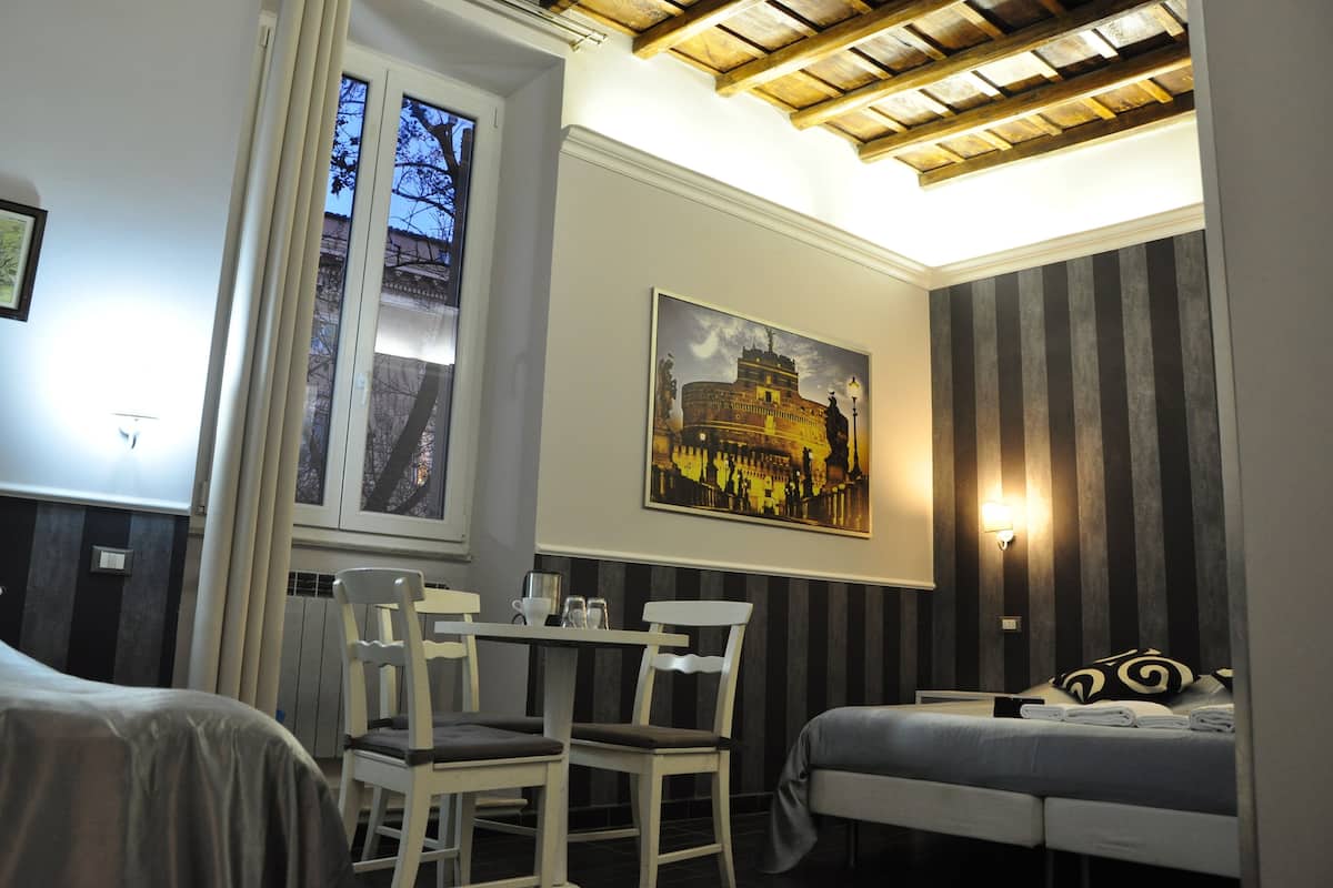 A hotel room with a bed, a dining table with chairs, a window with curtains, a painting on the wall, and a striped wall panel.