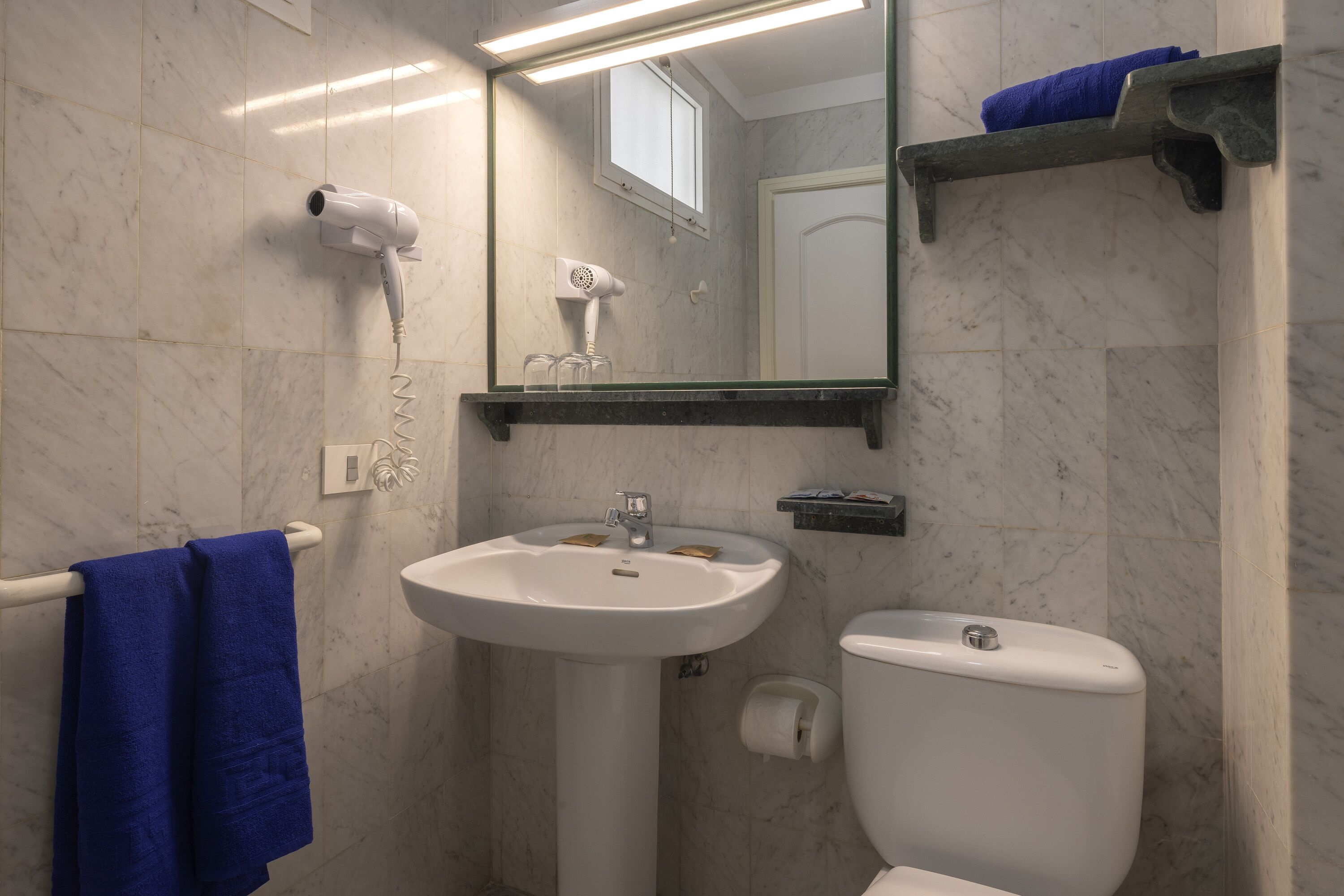 Un baño con lavamanos blanco, inodoro, secador de pelo y toalla azul en un perchero.