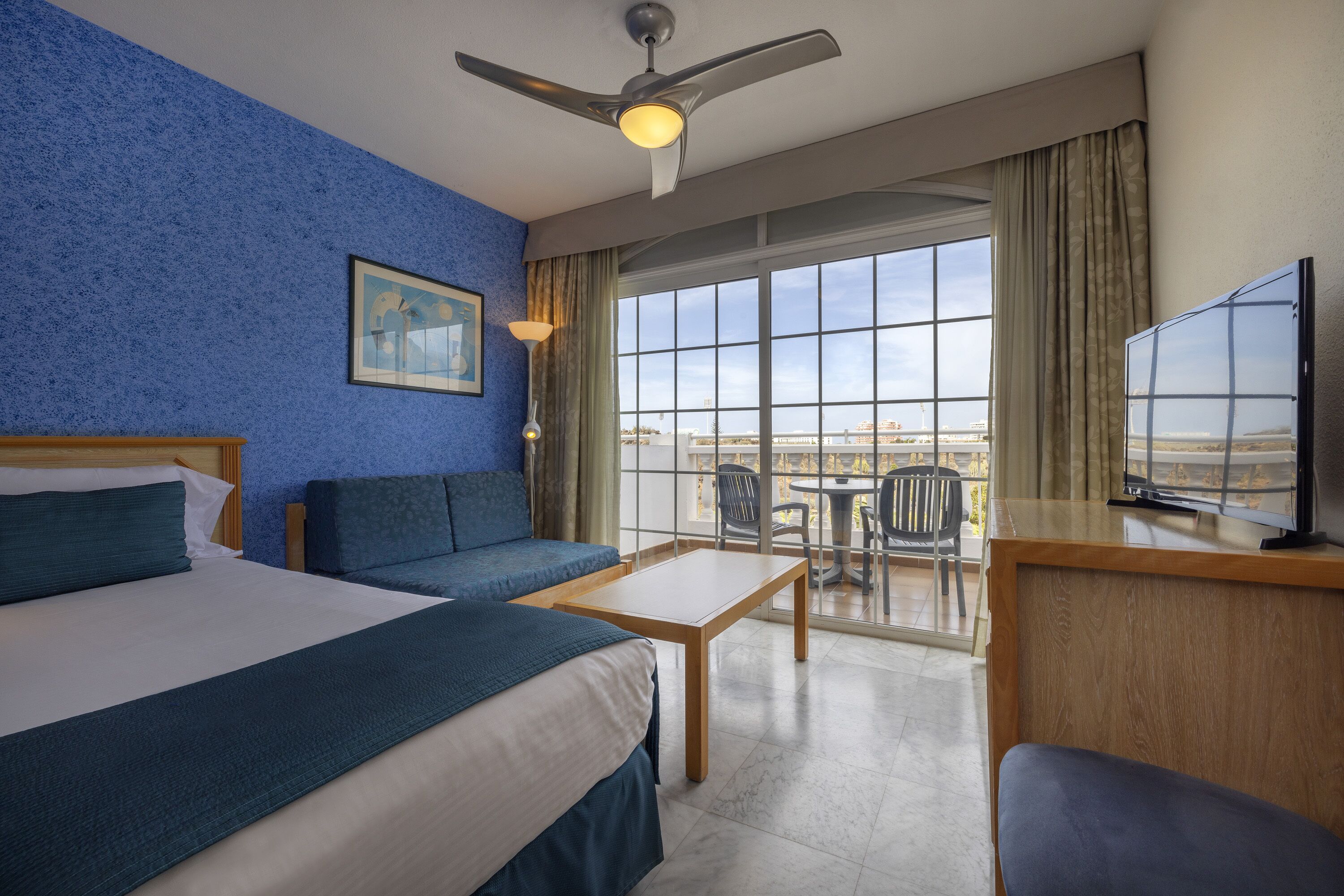 A hotel room with a bed, a sofa, a desk, a TV, and a balcony with a view.