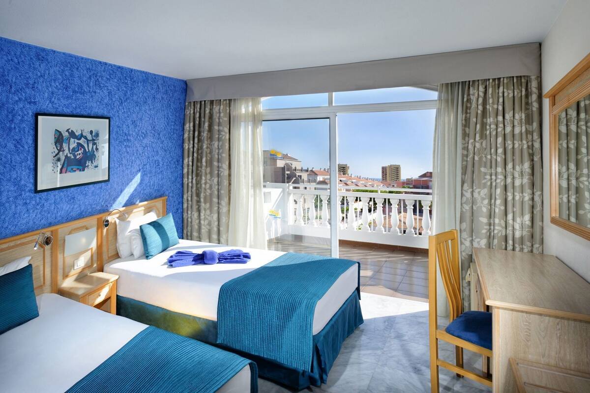 A hotel room with a bed, two bedside tables, a chair, a desk, a large window with a view, and a framed picture on the wall.