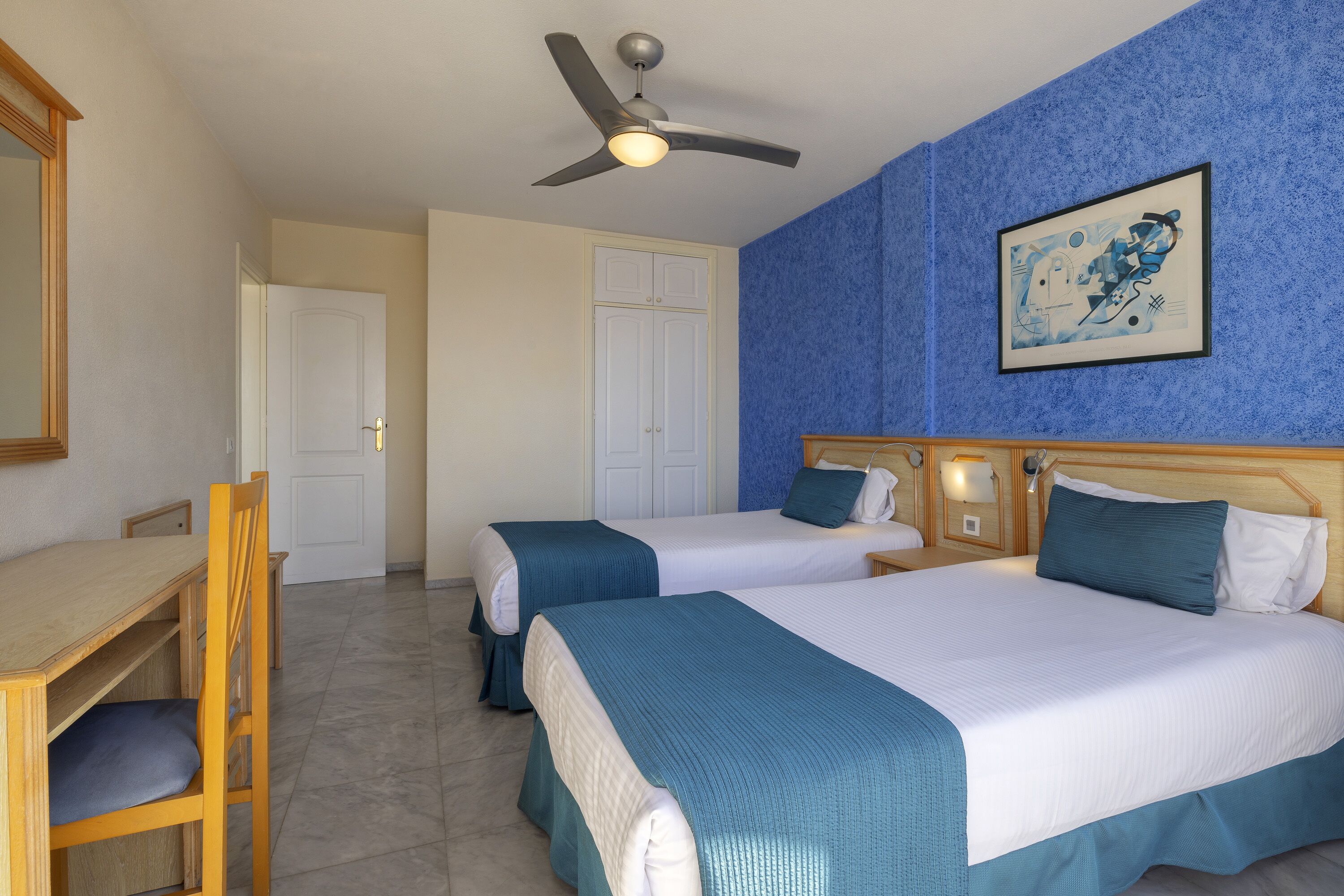 Une chambre d’hôtel avec deux lits, un bureau, une chaise, un ventilateur de plafond et une œuvre d’art encadrée au mur.