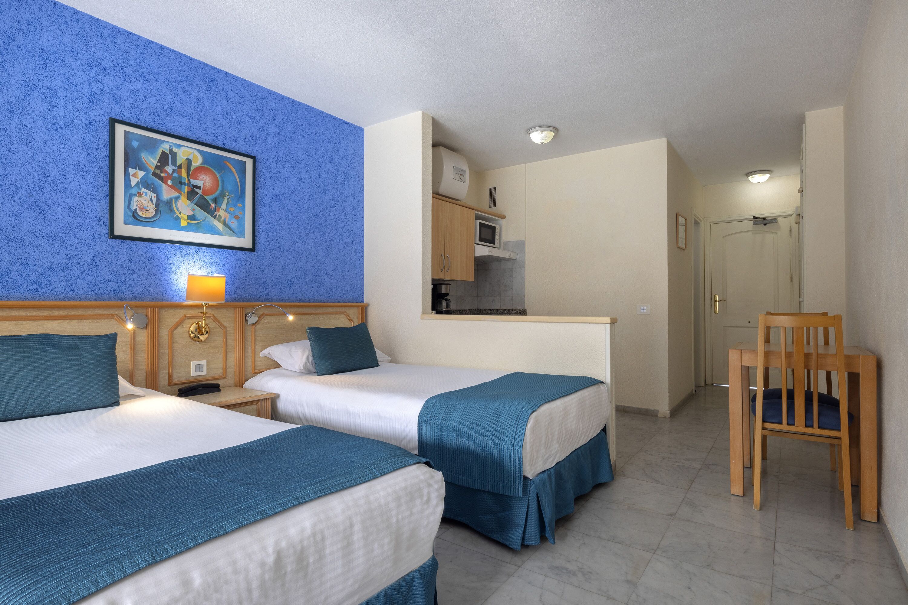 A hotel room with two beds, a painting on the wall, a desk with a lamp, a microwave, and a chair.