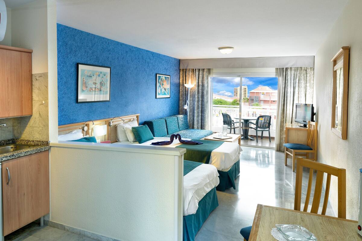 A hotel room with a kitchenette, a bed with a view, a dining area, and a balcony with a TV.