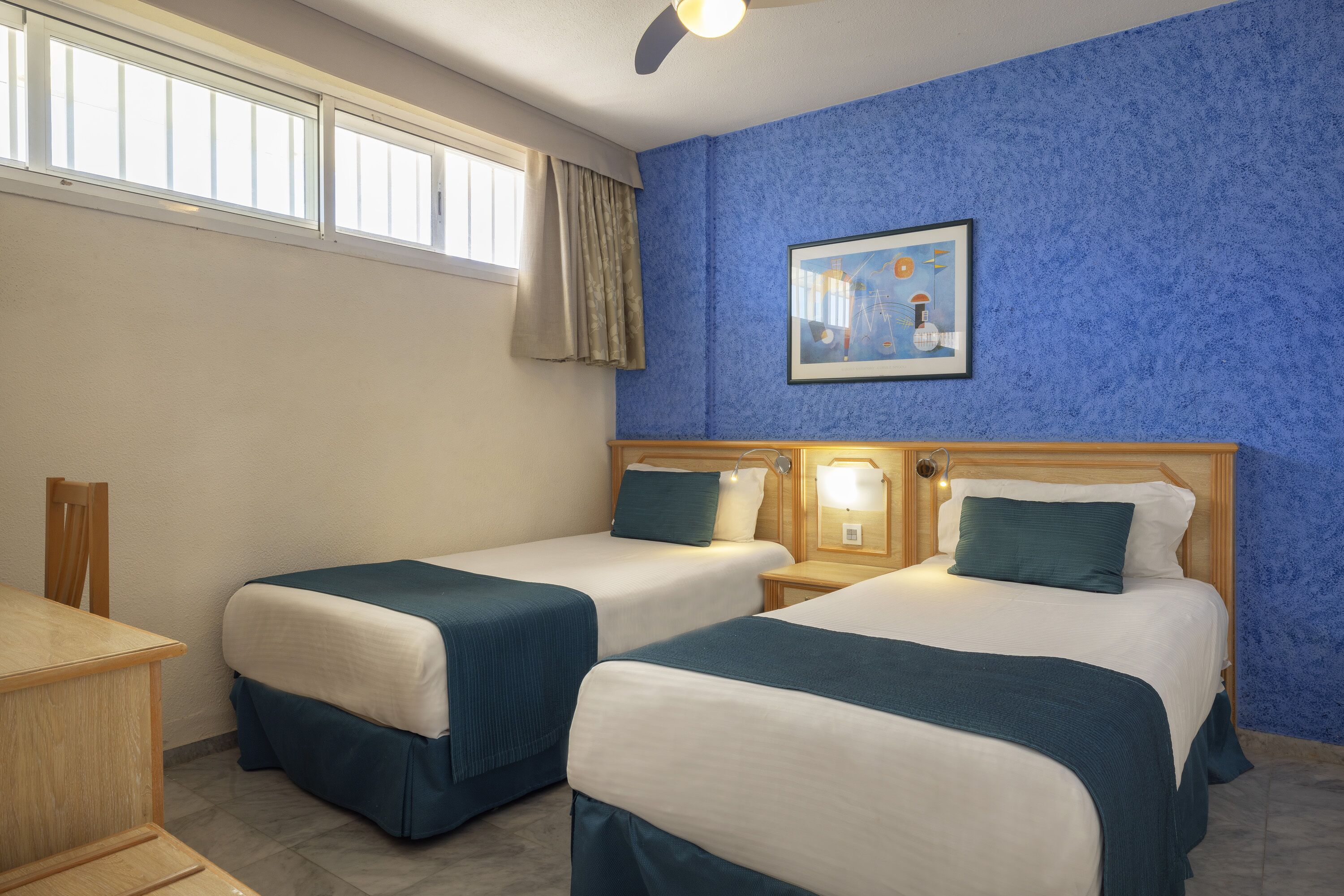 Habitación de hotel con dos camas, un escritorio, una silla, ventilador de techo, ventana con cortinas y un cuadro en la pared.
