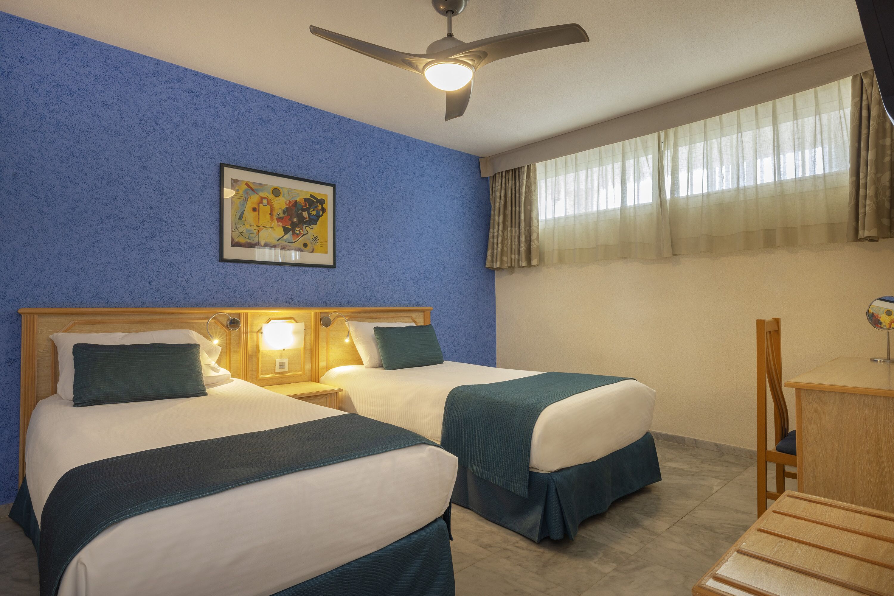 Una habitación de hotel con dos camas, un ventilador de techo, un cuadro en la pared y una ventana con cortinas.