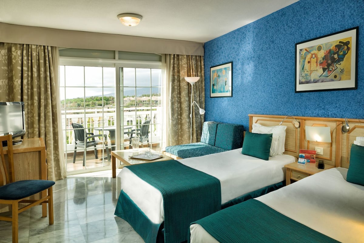 A hotel room with two beds, a desk with a chair, a TV, and a balcony with a view.
