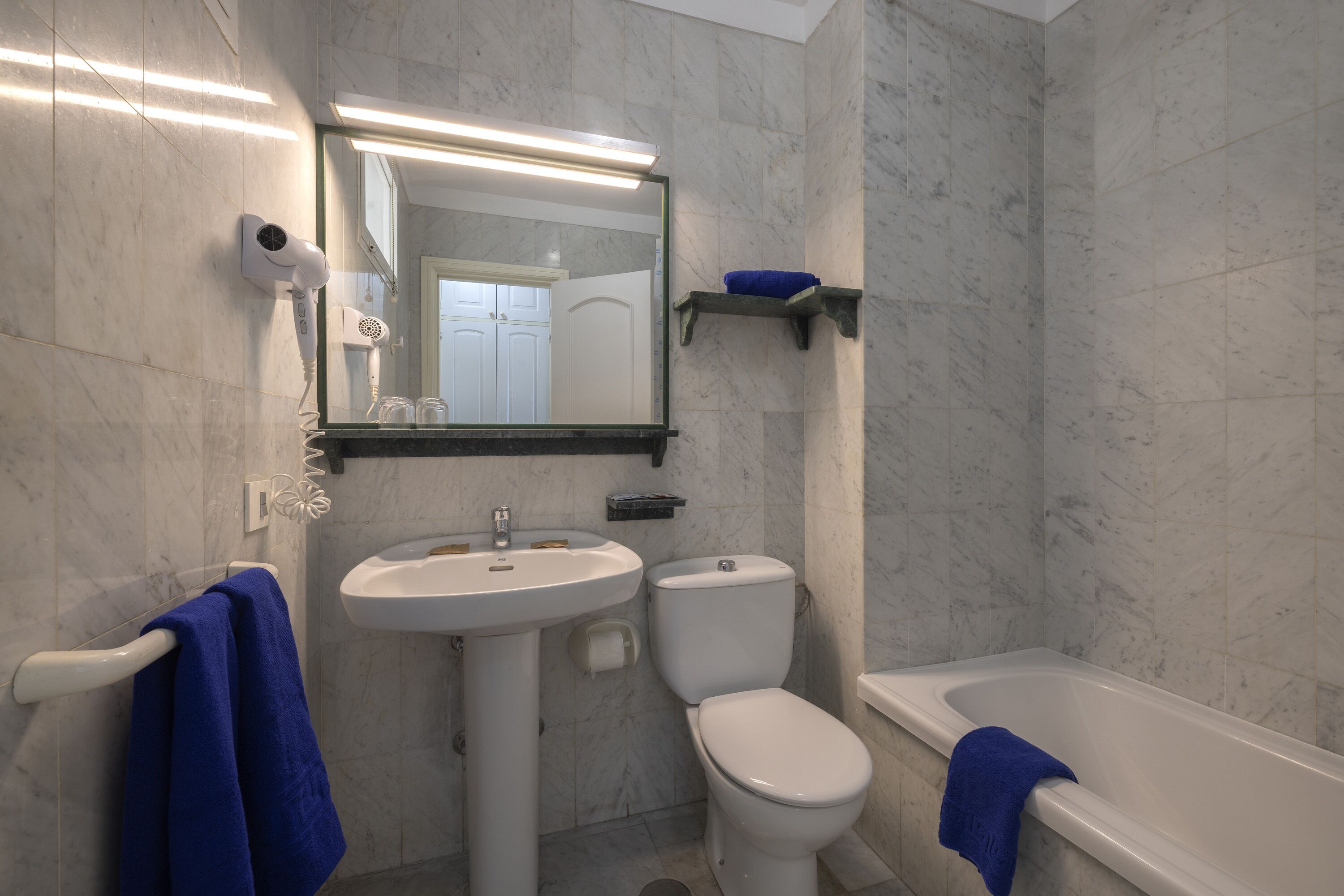 A bathroom with a white sink, toilet, and bathtub, marble walls, and a towel rack with blue towels.