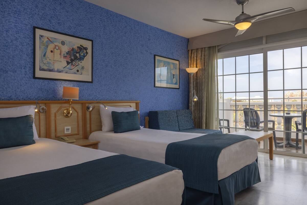 A hotel room with two beds, a ceiling fan, large windows, and artwork on the walls.