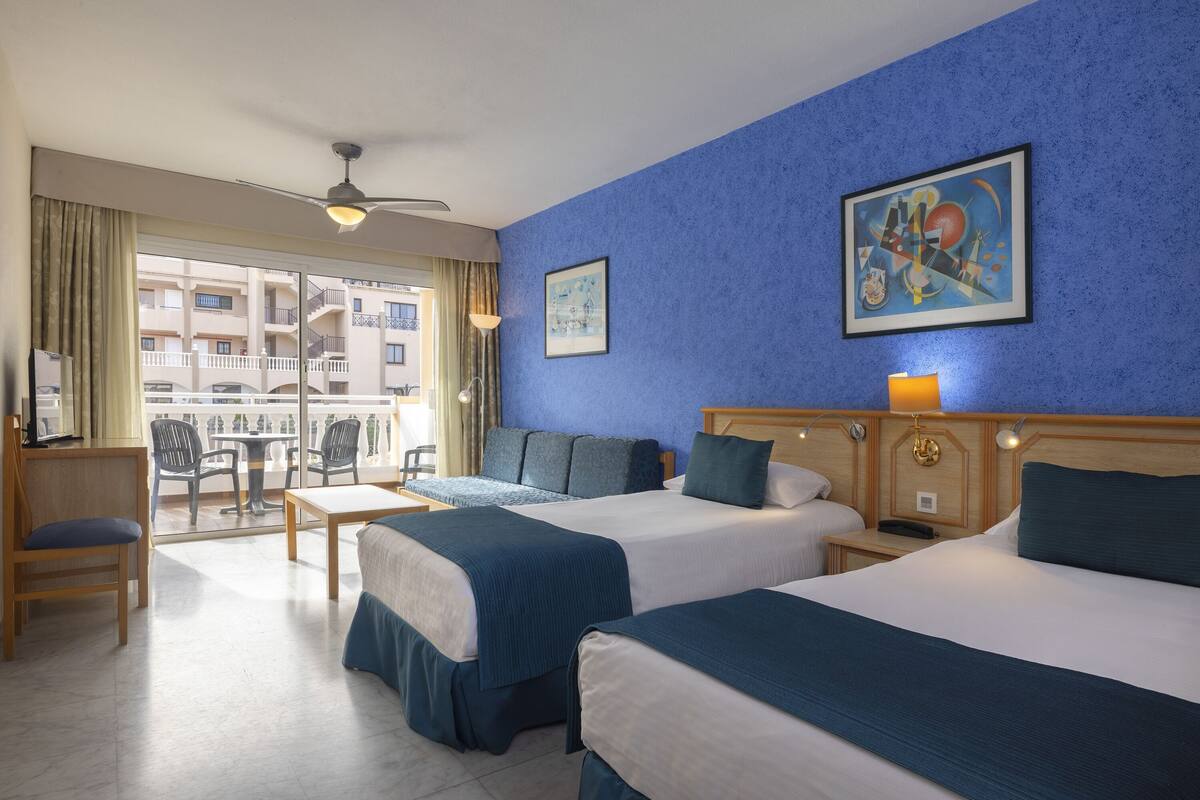 A hotel room with two beds, a desk, and a balcony with a view of the city.