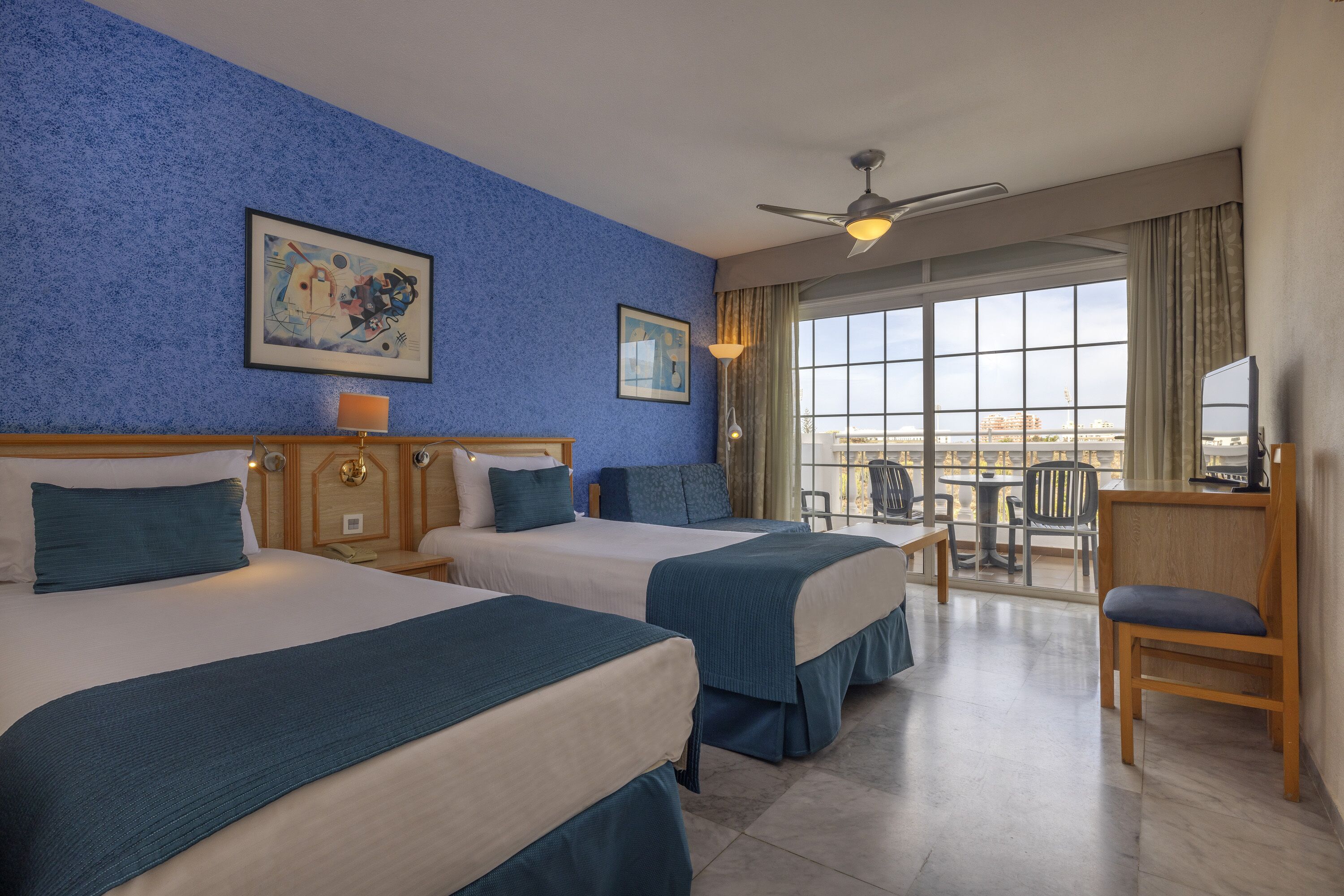 Habitación de hotel con dos camas, un escritorio, una silla, un ventanal con vistas y un ventilador de techo.