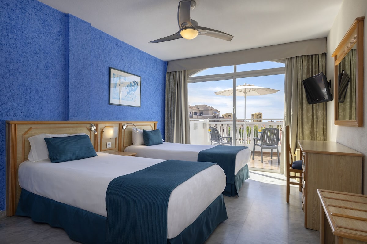 A hotel room with two beds, a ceiling fan, a large window with curtains, a desk, and a TV.