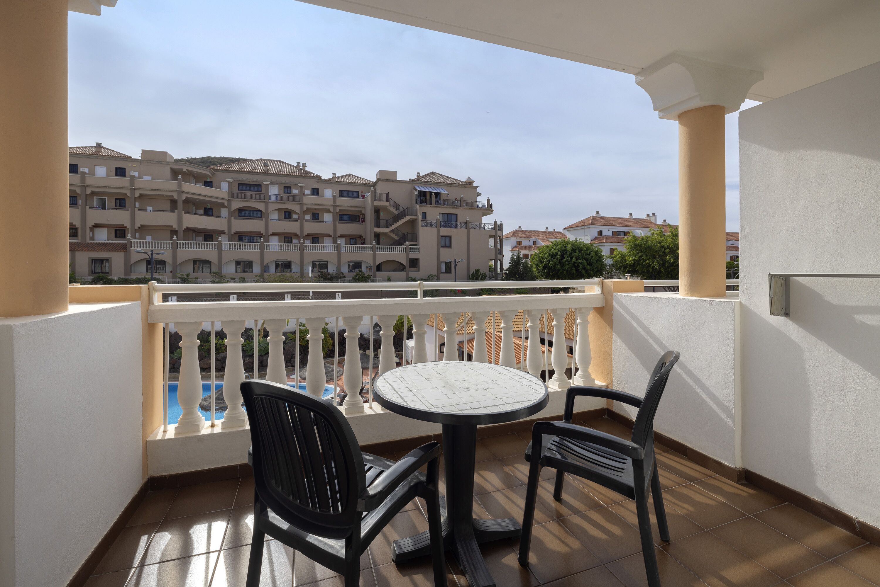 Un balcon doté d’une table ronde et de deux chaises, offrant une vue sur un immeuble à multiples balcons et une piscine.