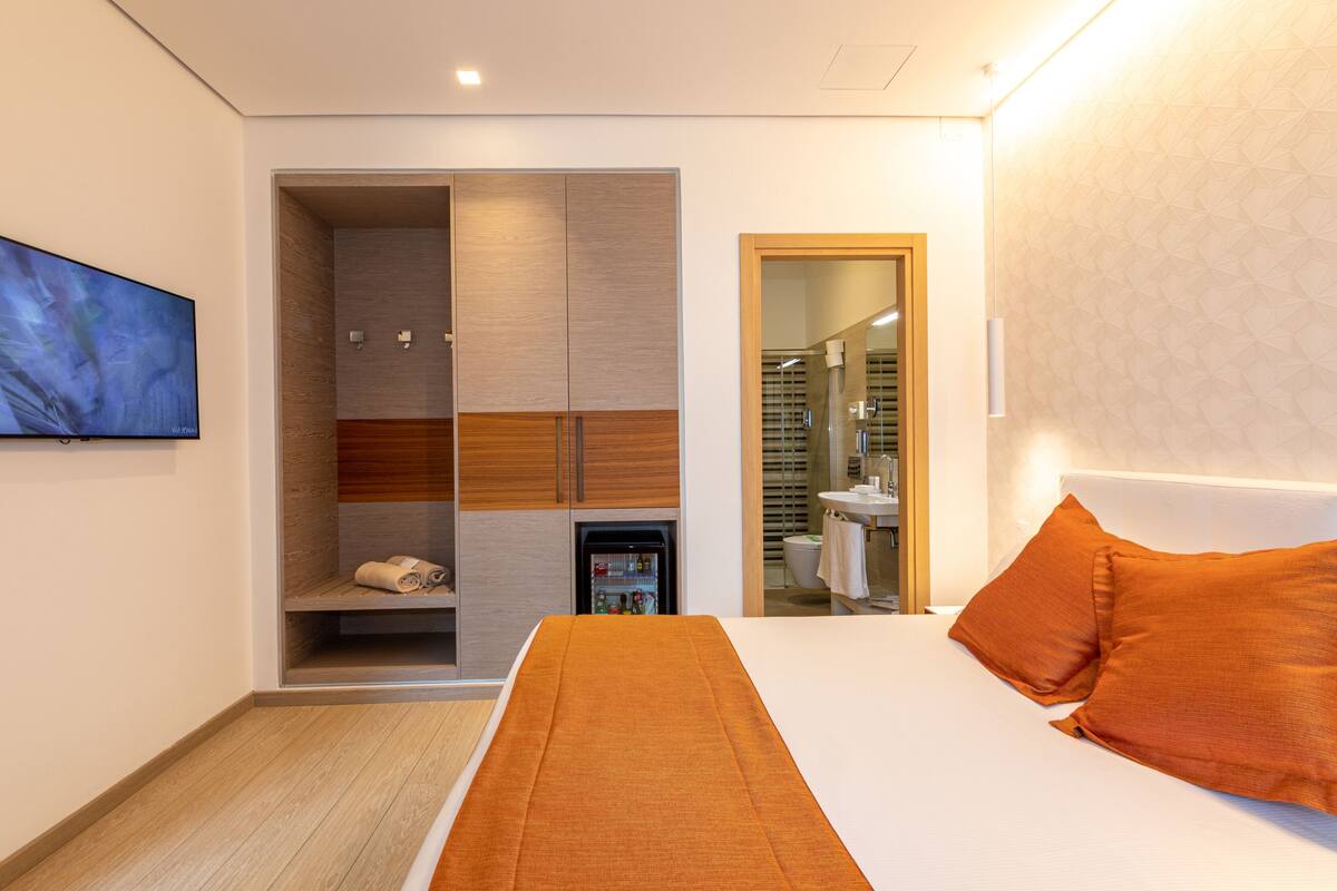 A modern hotel room with a bed, a built-in wardrobe, a small refrigerator, and a bathroom visible through an open door.