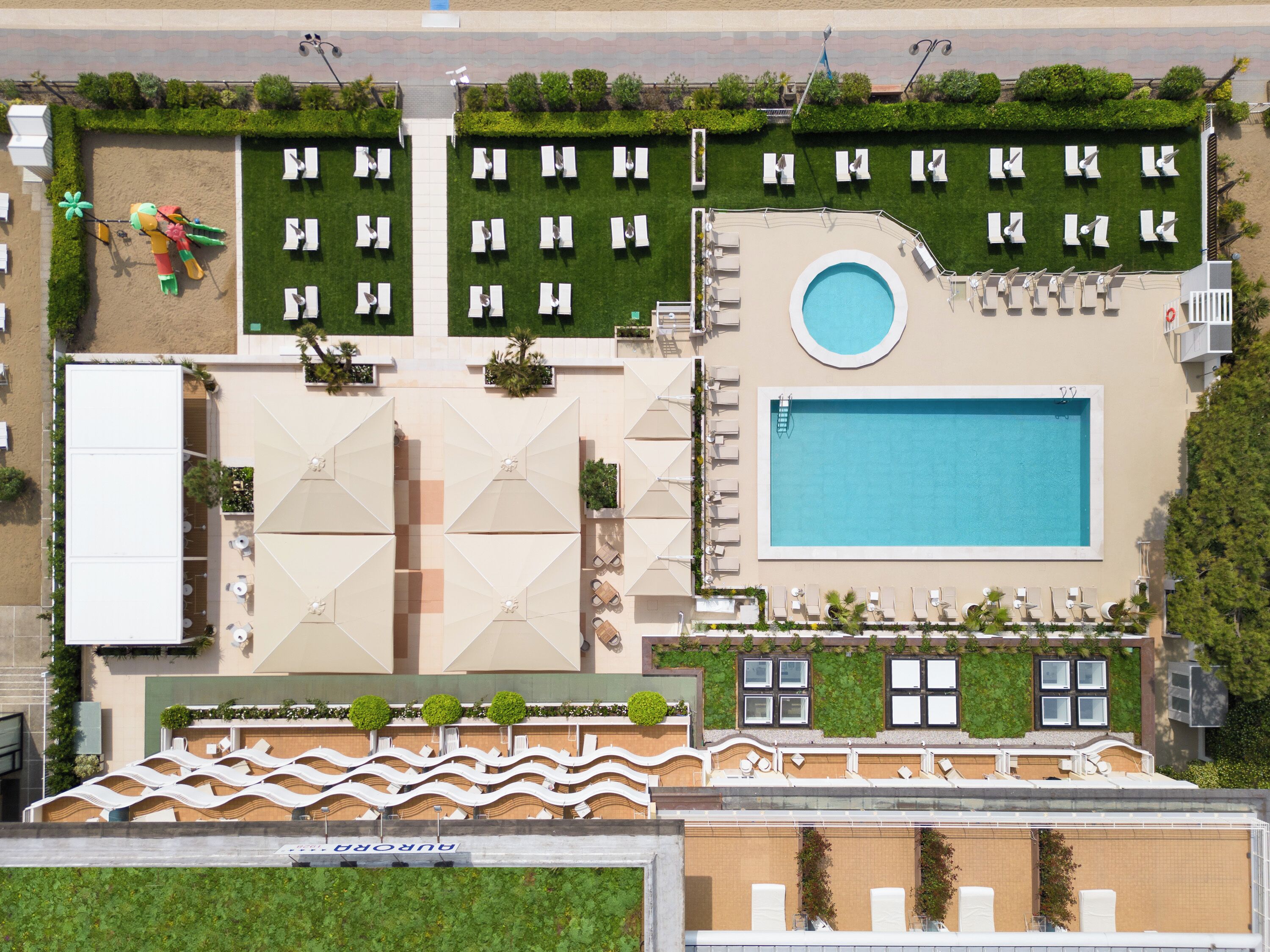An aerial view of a hotel with a swimming pool, multiple lounge areas with sunbeds, and a well-maintained lawn.