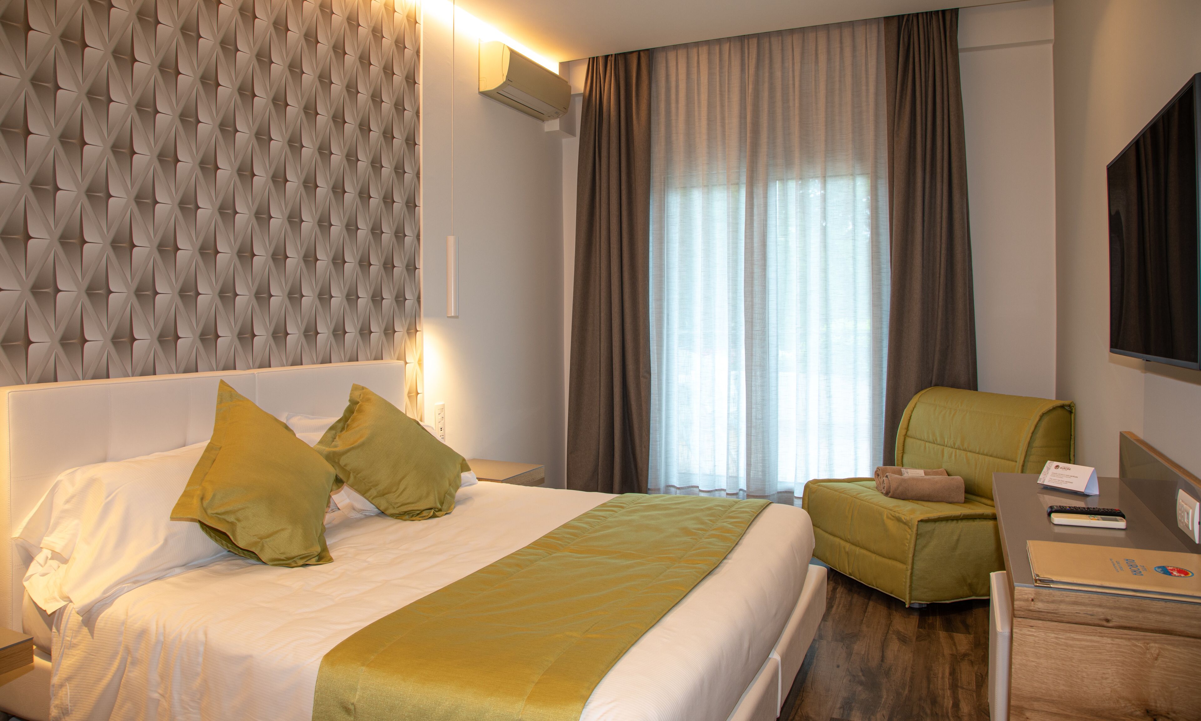 A hotel room with a bed, a green armchair, a desk with a computer, and a large window with curtains.