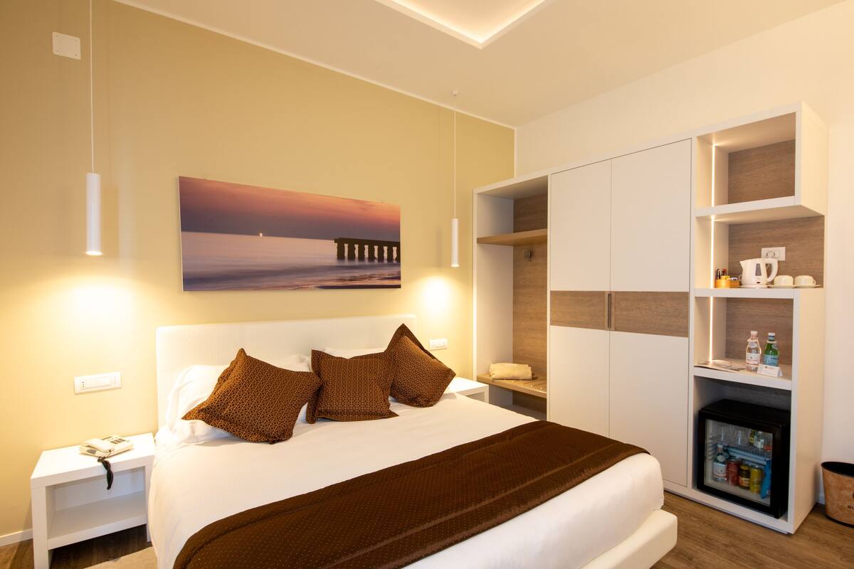 A modern hotel room with a large bed, a painting of a sunset over the sea, a built-in wardrobe, and a minibar.