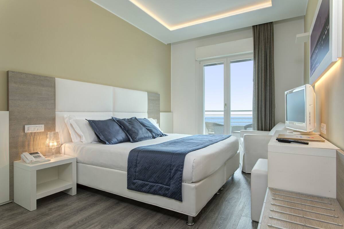 A modern hotel room with a large bed, bedside tables, a TV, and a view of the sea.