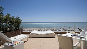 A terrace with a white sofa, a table with a bottle, glasses, and a magazine, overlooking a beach with boats and a clear sky.