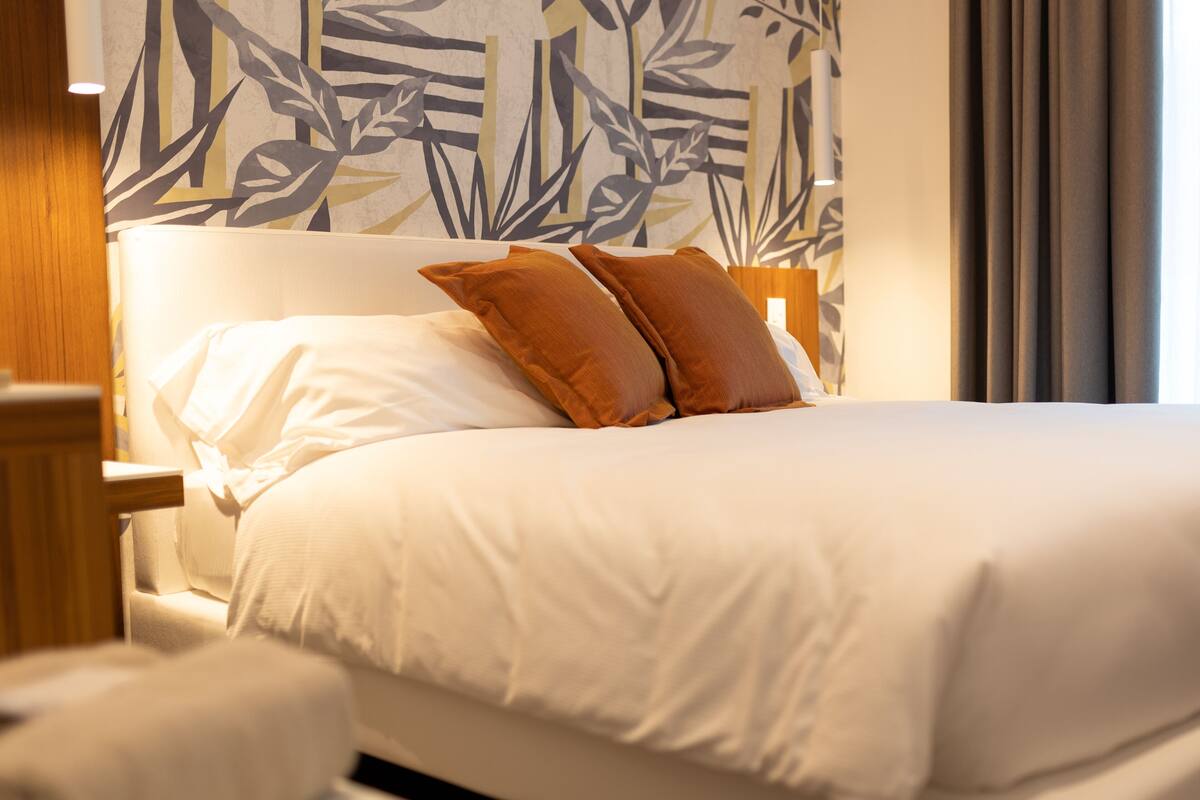 A neatly made bed with white linens and two brown pillows, a wooden headboard, and a wall with a leafy pattern.
