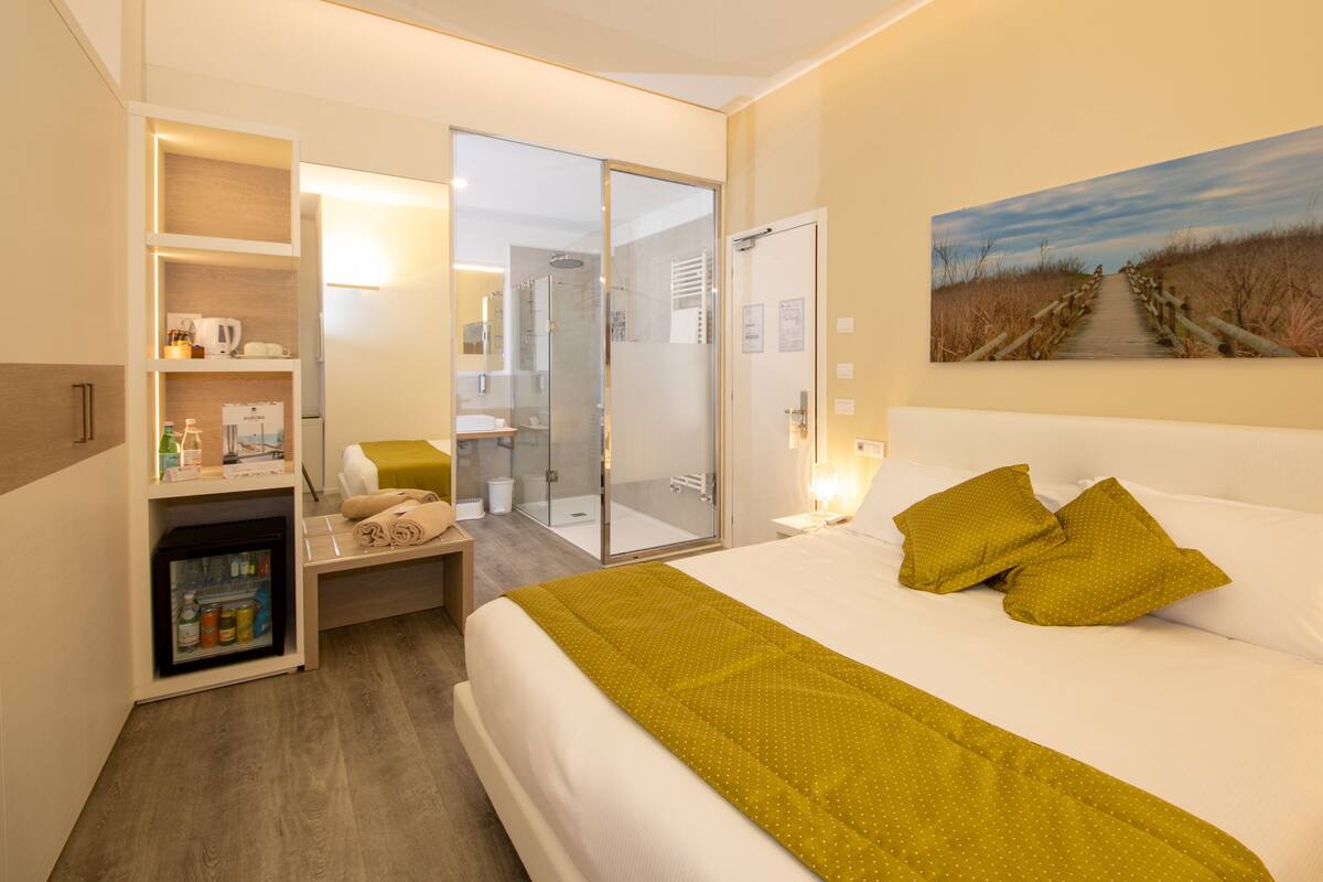 A hotel room with a bed, a small seating area, a minibar, and a glass shower enclosure.