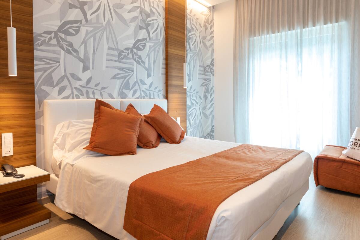 A modern hotel room with a large bed, a bedside table with a phone, and a small seating area with a cushion.