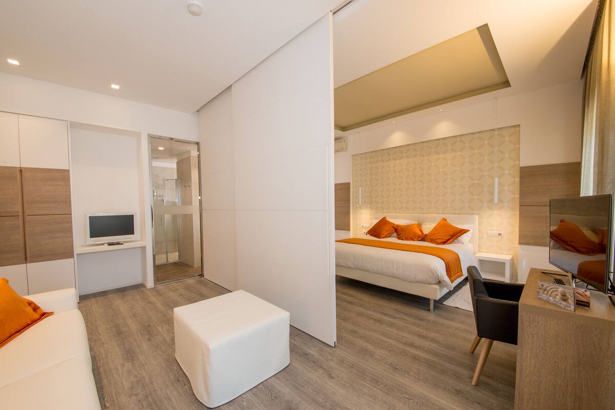 A modern hotel room with a bed, a sofa, a TV, and a desk.