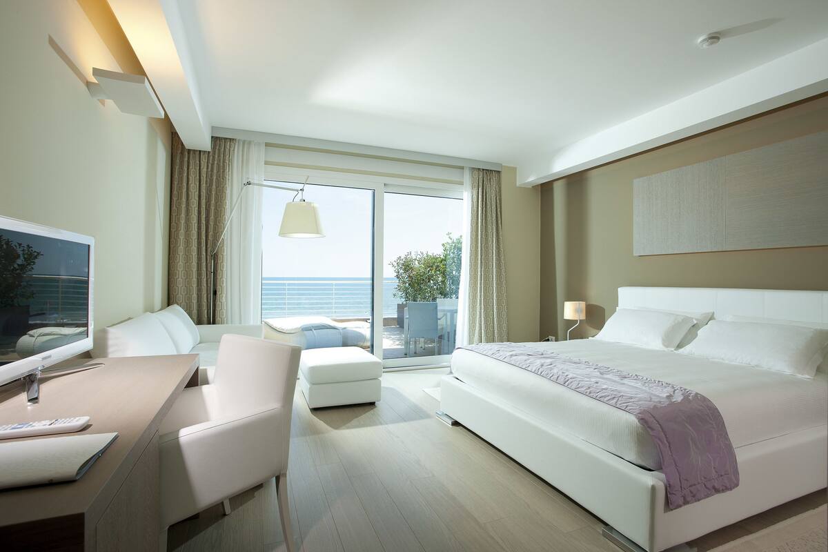 A modern hotel room with a bed, a desk with a computer, a chair, and a view of the outdoors.