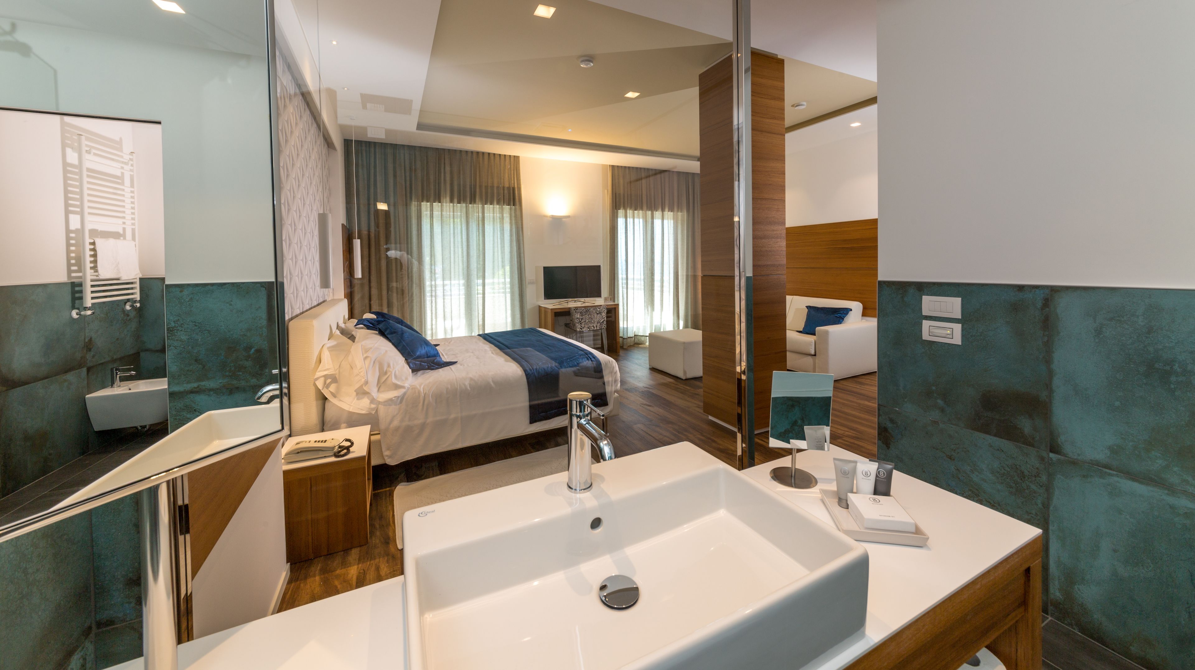 Una habitación de hotel moderna con una cama grande, un baño con lavamanos y espejo, y una zona de estar con escritorio y silla.