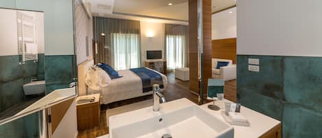 Una habitación de hotel moderna con una cama grande, un baño con lavamanos y espejo, y una zona de estar con escritorio y silla.