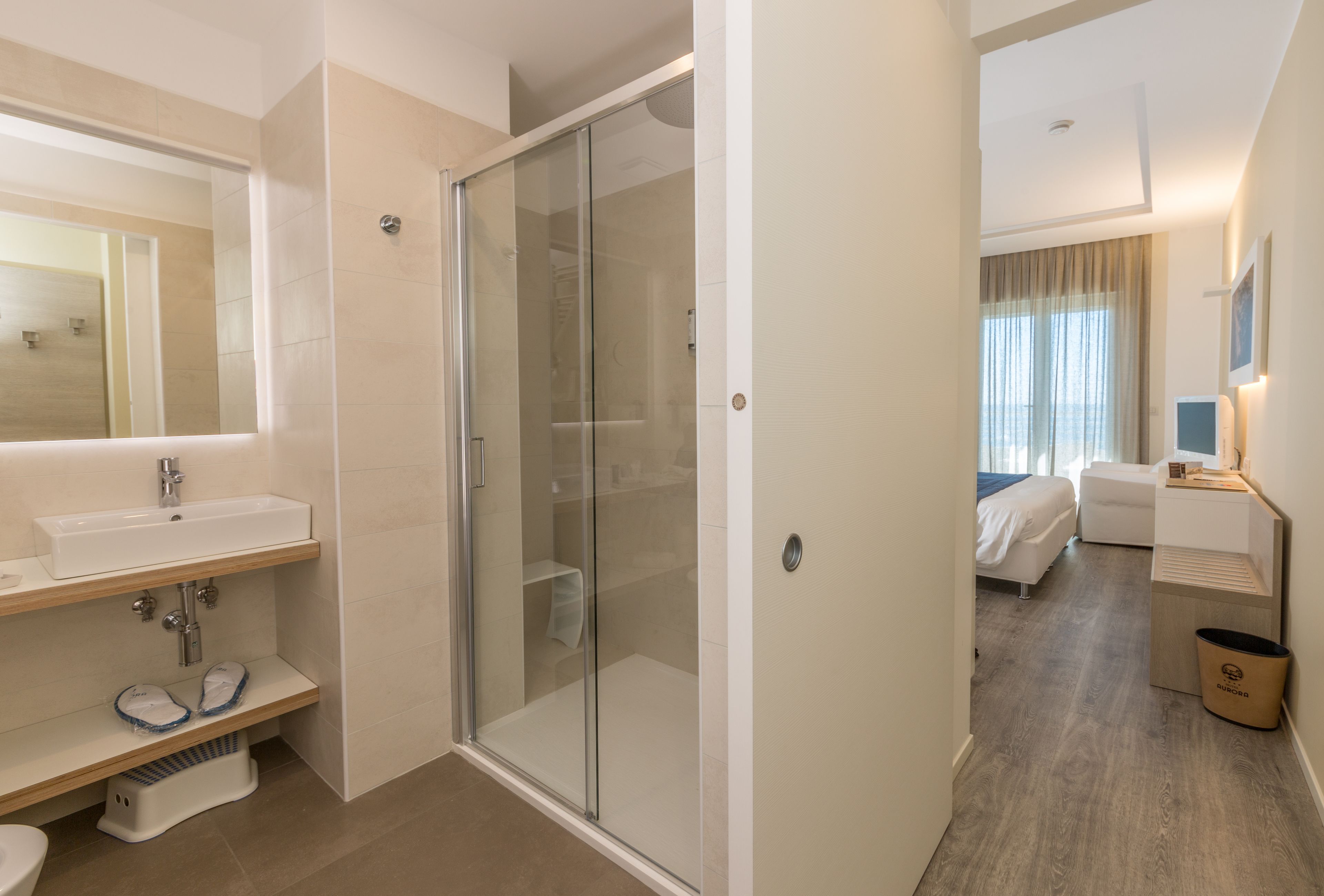 Une salle de bain d’hôtel moderne avec une douche en verre, un lavabo avec un miroir et une table de chevet avec une lampe.