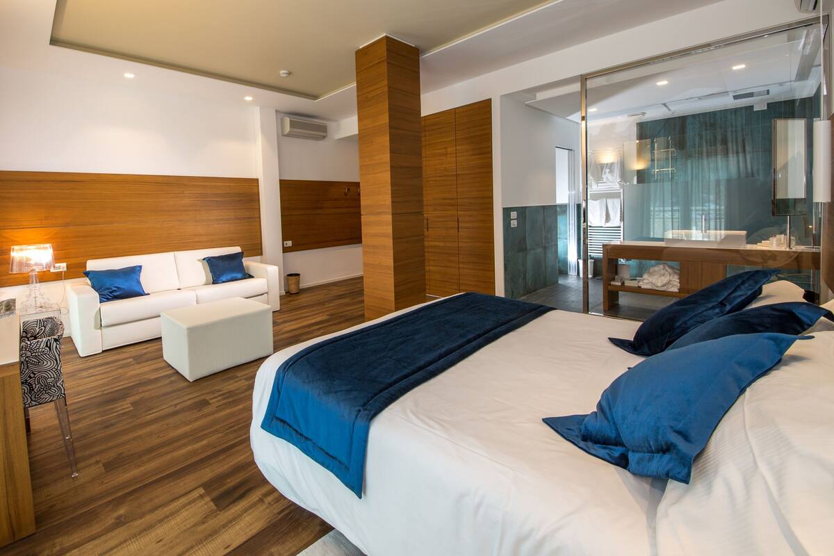 A modern hotel room with a large bed, a sitting area with a sofa and armchairs, a desk with a computer, and a glass shower enclosure.