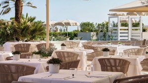 An outdoor dining area with wicker chairs and tables, covered by umbrellas, and surrounded by greenery.