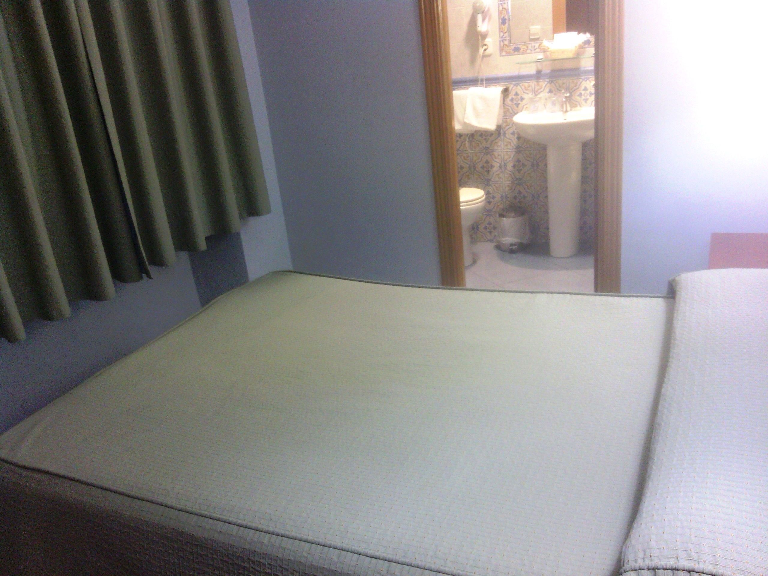 A room with a bed, a mirror reflecting a bathroom, and a window with curtains.