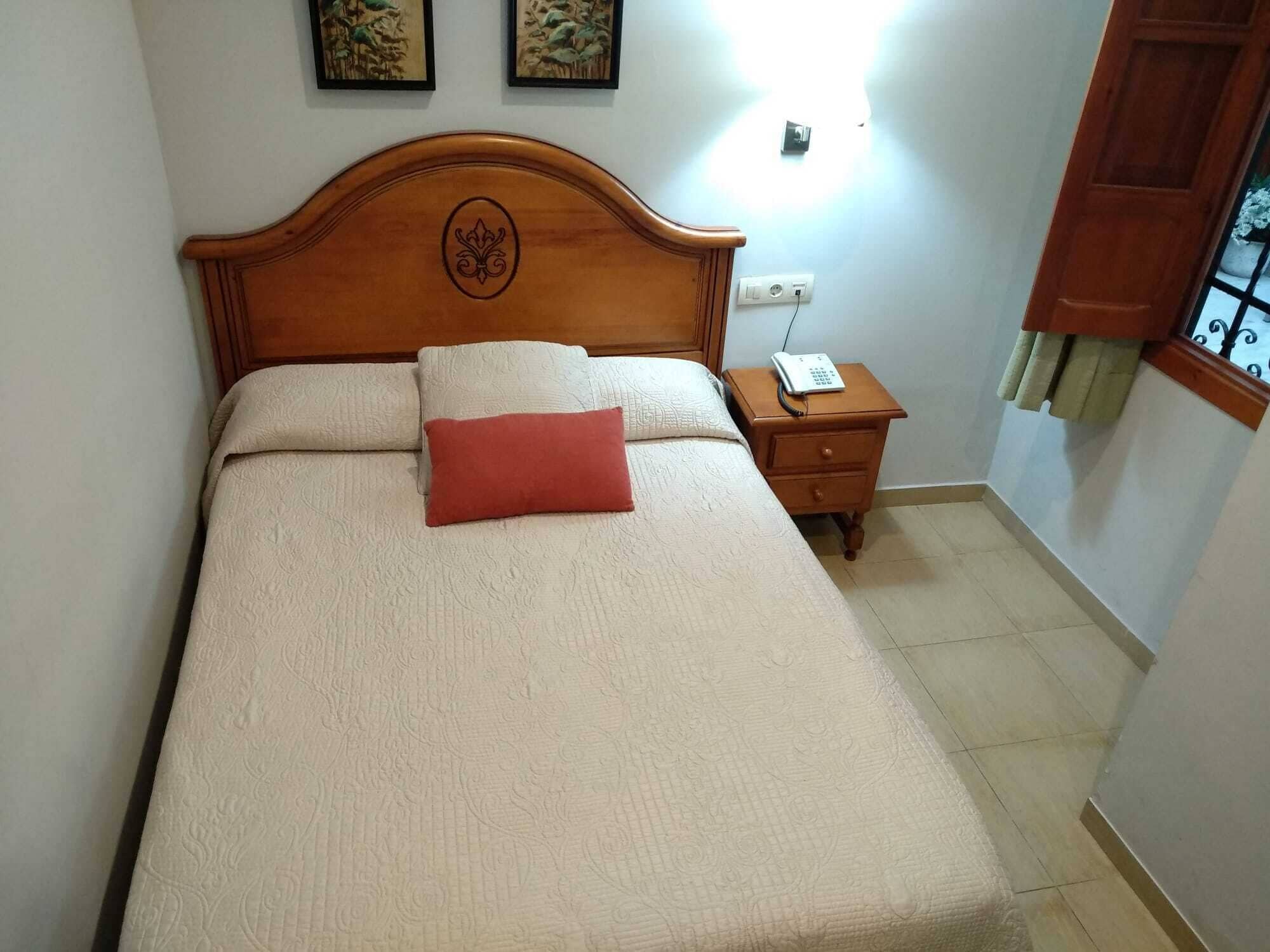 A neatly made bed with a wooden headboard, a red pillow, and a bedside table with a phone.